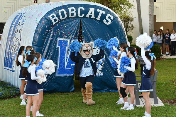 High school cheerleaders and mascot in front of an inflatable tunnel with 'BOBCATS' text at a football game