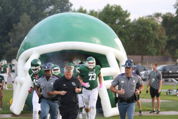 Football team and coaches running onto the field through a green and white inflatable helmet tunnel, flanked by state troopers