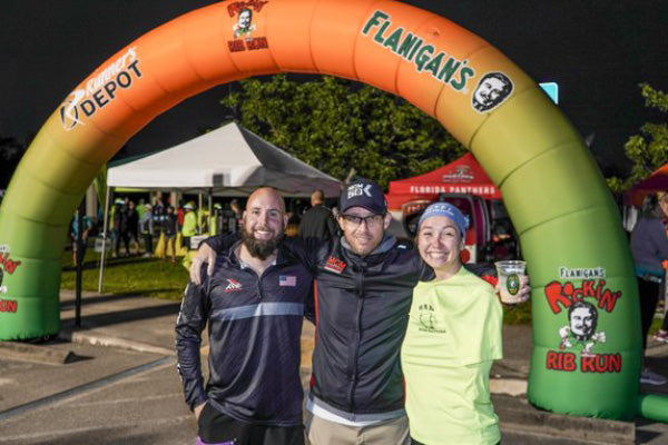 Three runners smiling at a race event in front of an orange inflatable arch sponsored by Flanigan's and Home Depot