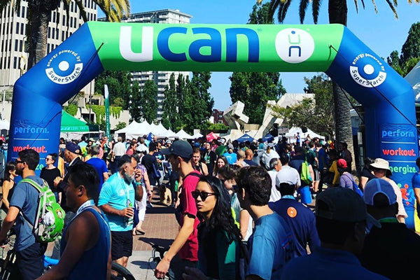 Event participants gathering under a blue and green UCAN branded inflatable arch at an outdoor fitness expo