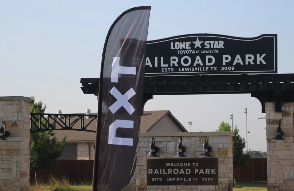 Feather flag with the logo NXT situated next to the prominent stone archway of Railroad Park.