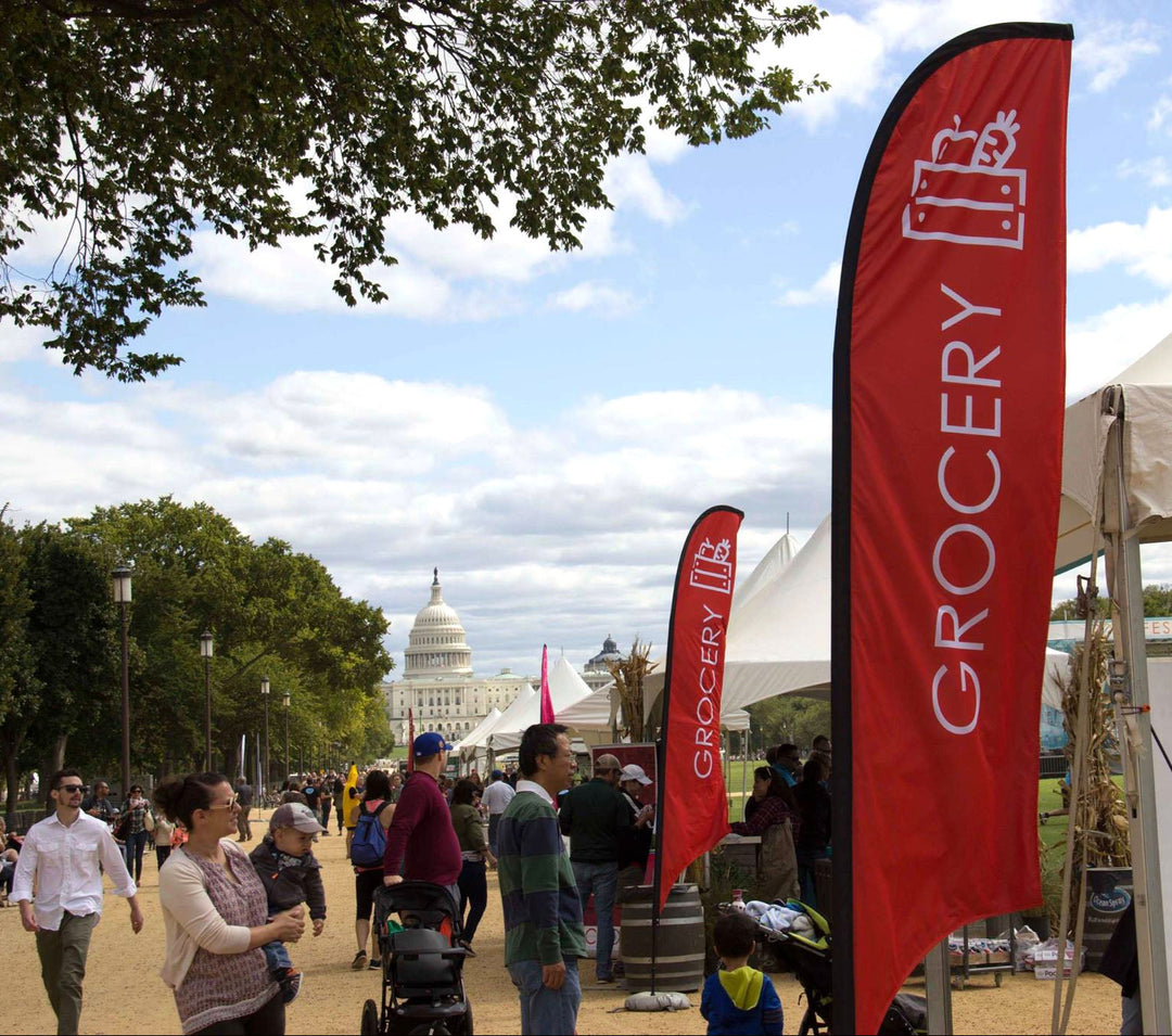 Red feather flag labeled “Grocery” at an outdoor event near the U.S. Capitol with white tents and people walking.