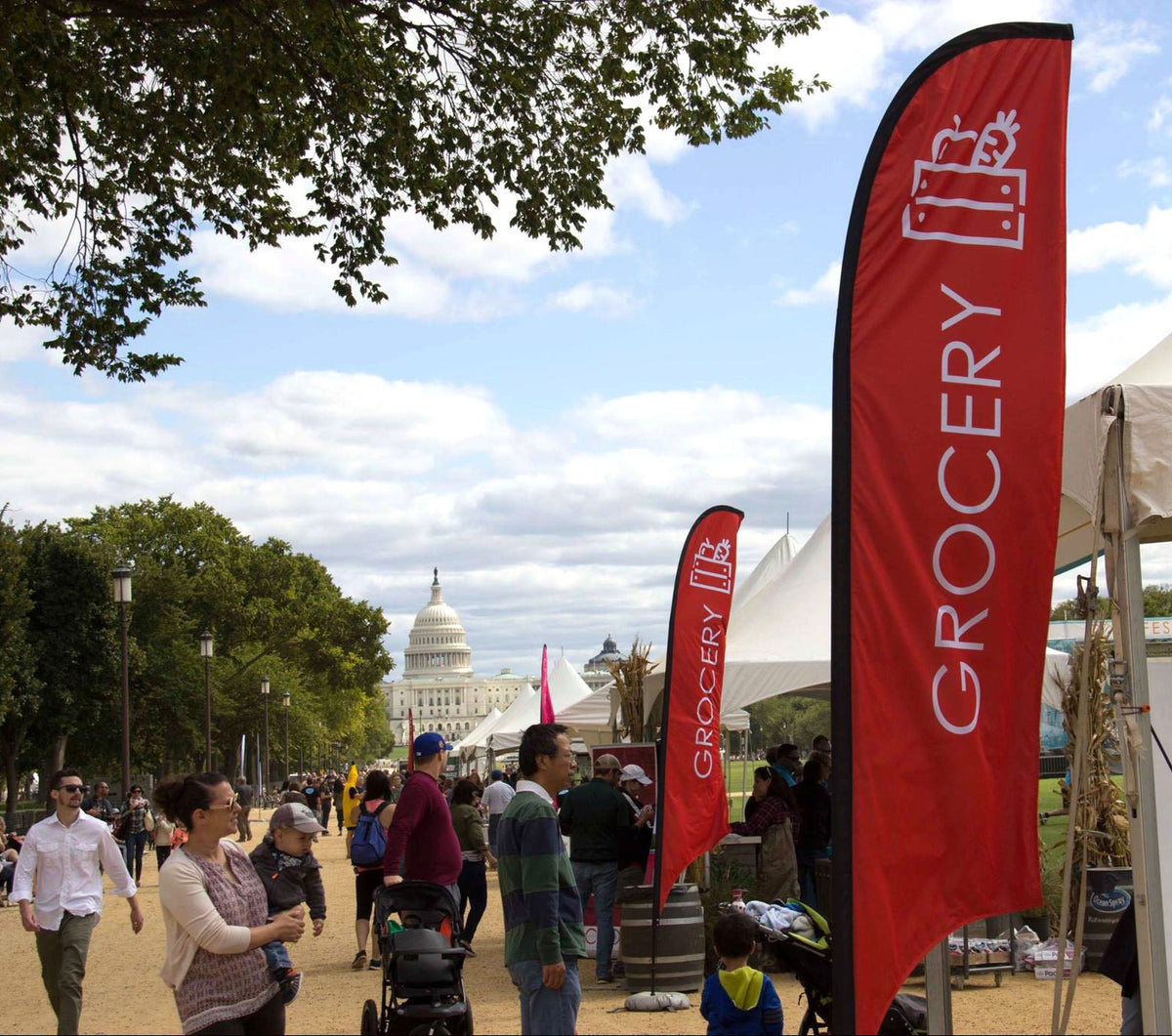 Red feather flag labeled “Grocery” at an outdoor event near the U.S. Capitol with white tents and people walking.