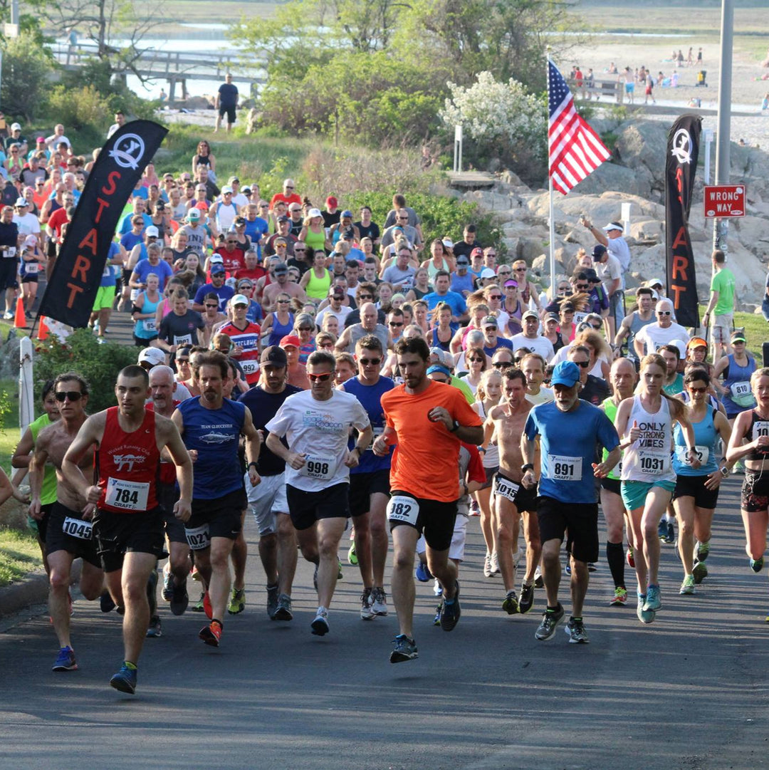 Large group of runners starting a road race beside black feather flags marked "START".