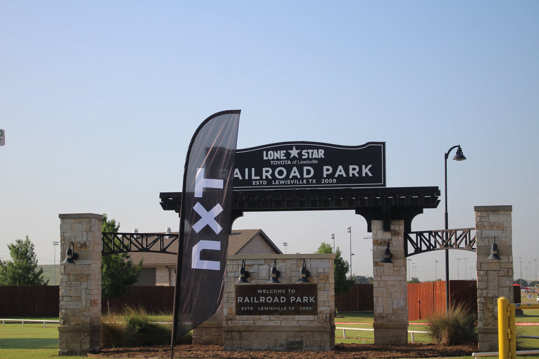 Black feather flag with white "NXT" text displayed in front of the Lone Star Toyota Railroad Park entrance in Lewisville, Texas