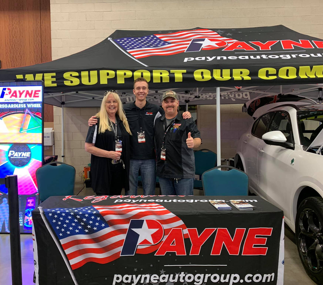 Payne Auto Group indoor event booth with staff posing in front of branded tent, American flag table cover, and prize wheel.