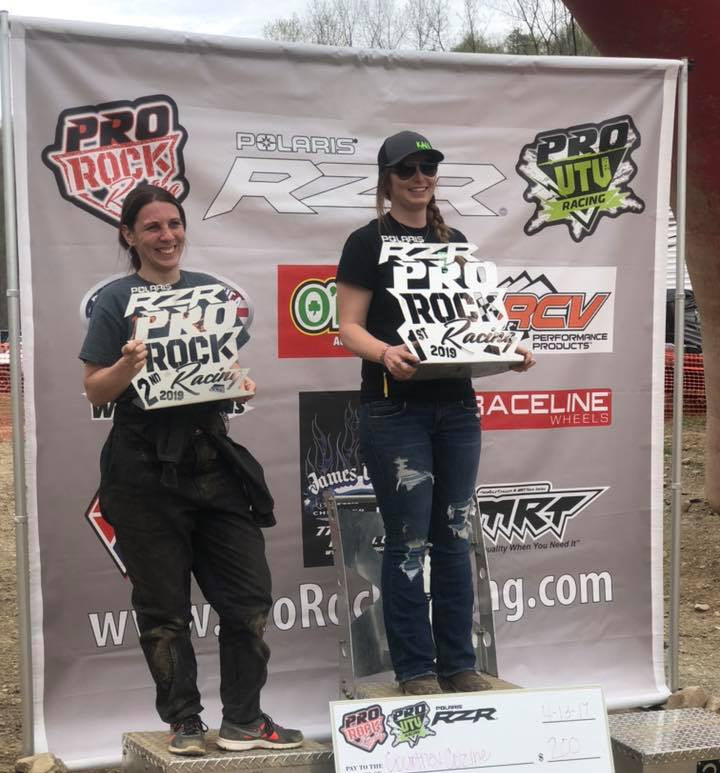 Two female off-road racers standing on a podium with trophies in front of a Pro Rock Racing vinyl banner featuring sponsor logos.