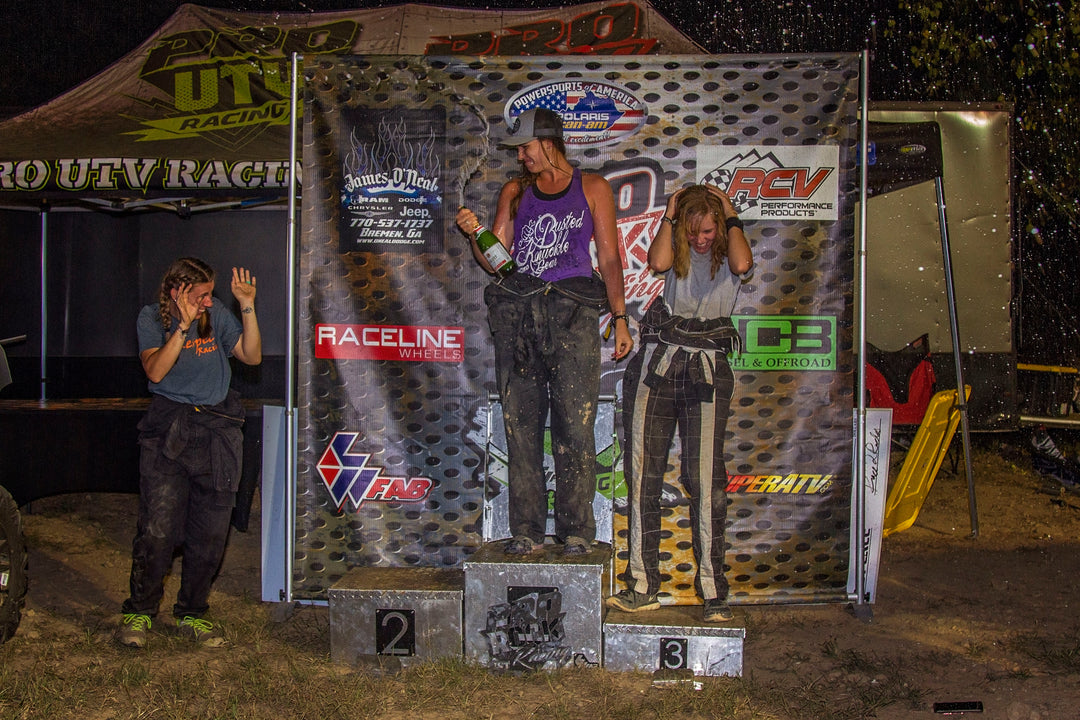 Female off-road race winners on a podium in front of a Pro Rock Racing vinyl banner, celebrating with champagne spray.