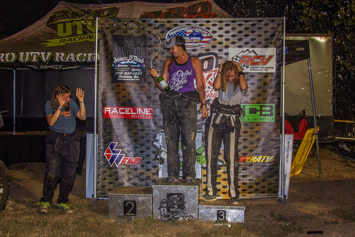 Female off-road race winners on a podium in front of a Pro Rock Racing vinyl banner, celebrating with champagne spray.