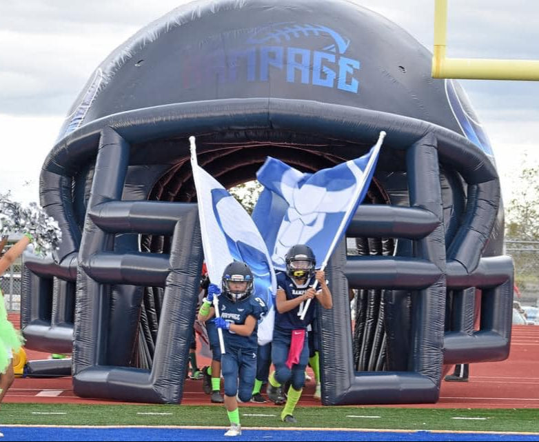 Kids in football uniforms run through inflatable "Rampage" helmet tunnel with flags