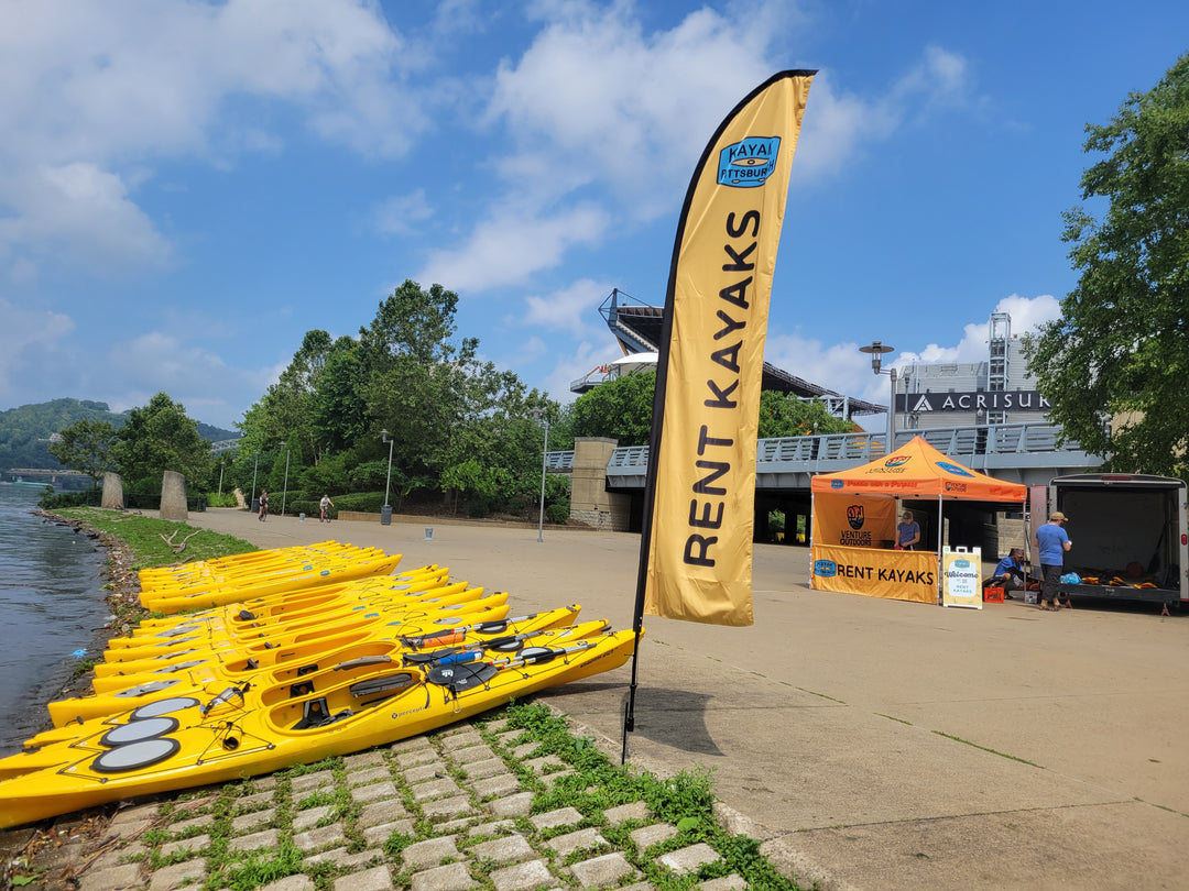 Yellow feather flag with "Rent Kayaks" printed on it, displayed near a kayak rental booth by the river in Pittsburgh