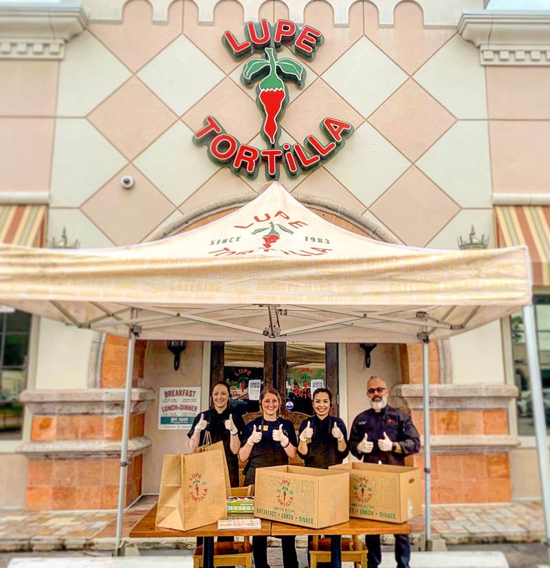 Cheerful team under a restaurant pop up tent at Lupe Tortilla, ready for outdoor service with boxes of freshly prepared food on display