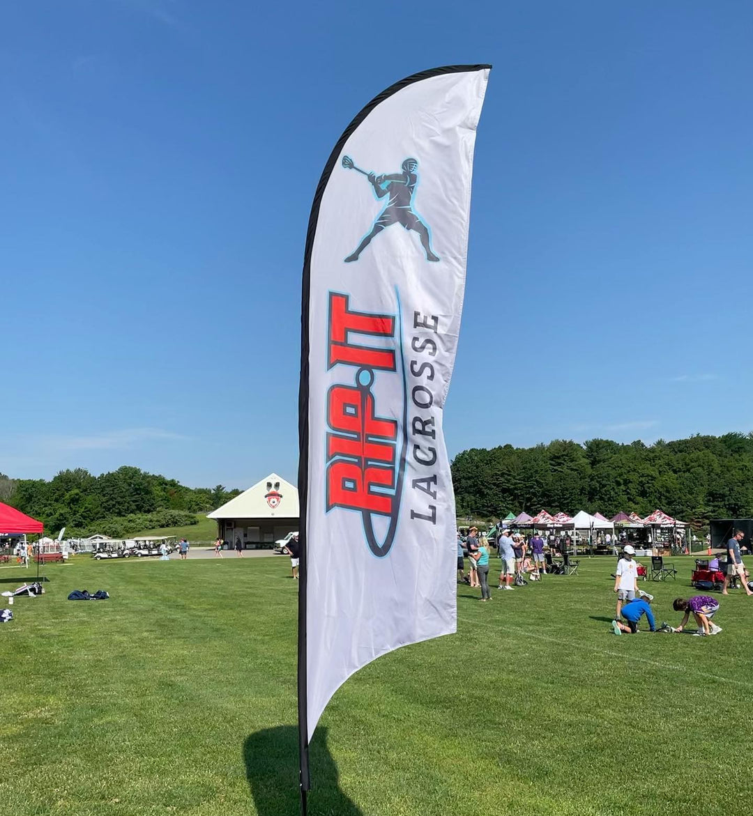 RIP-IT Lacrosse feather flag standing on a green field during a youth sports tournament under clear blue skies.