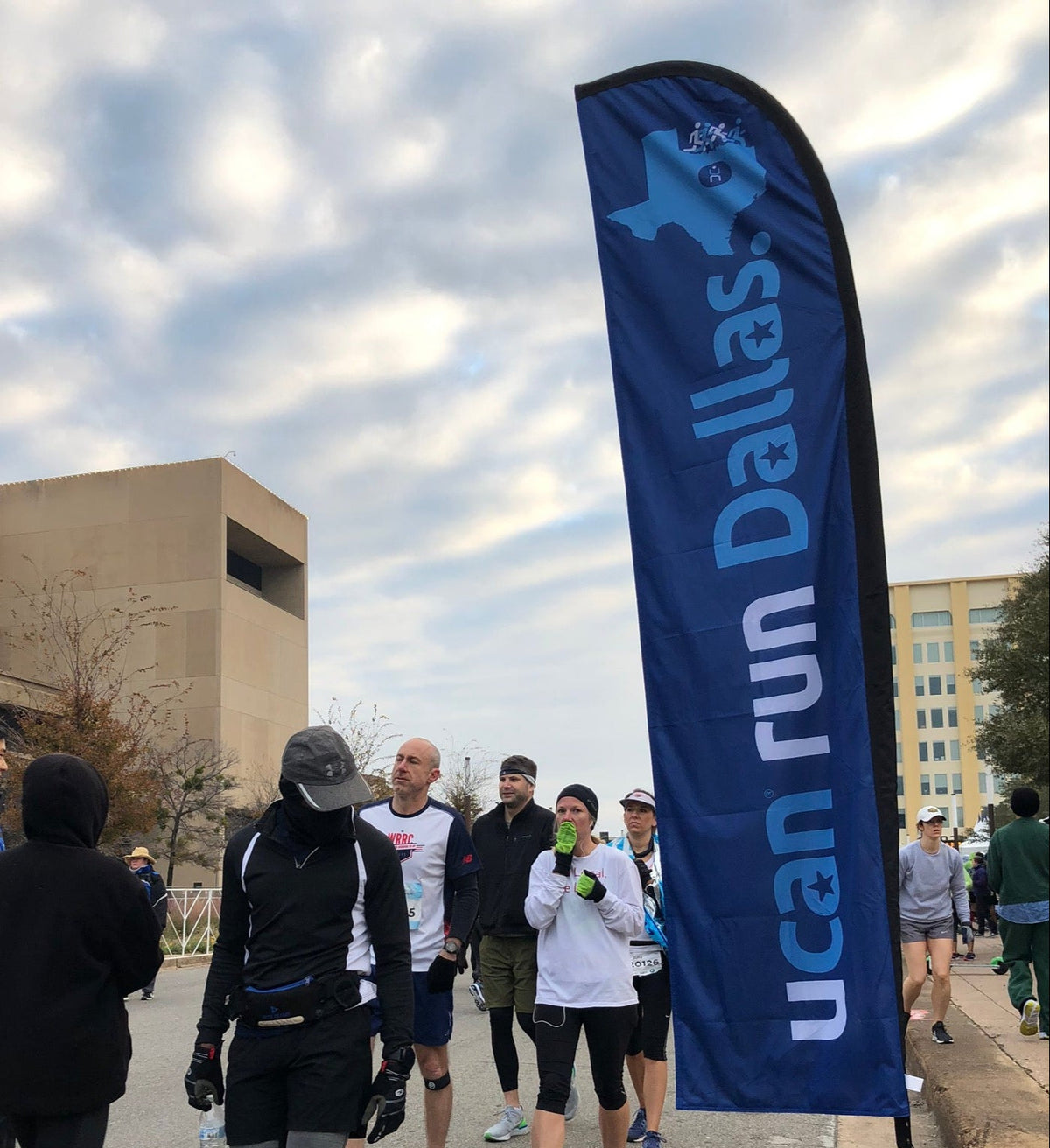 Group of runners passing by a vertical UCAN "Run Dallas" feather flag at a race event in Dallas.