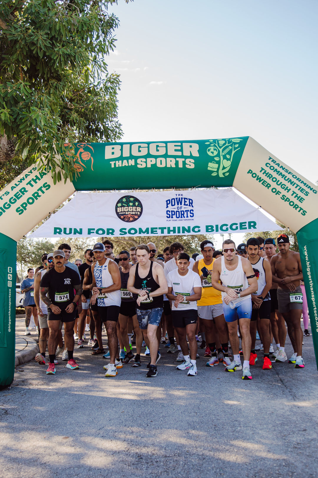 Crowd of runners preparing to start race under a green and white "Bigger Than Sports" inflatable archway