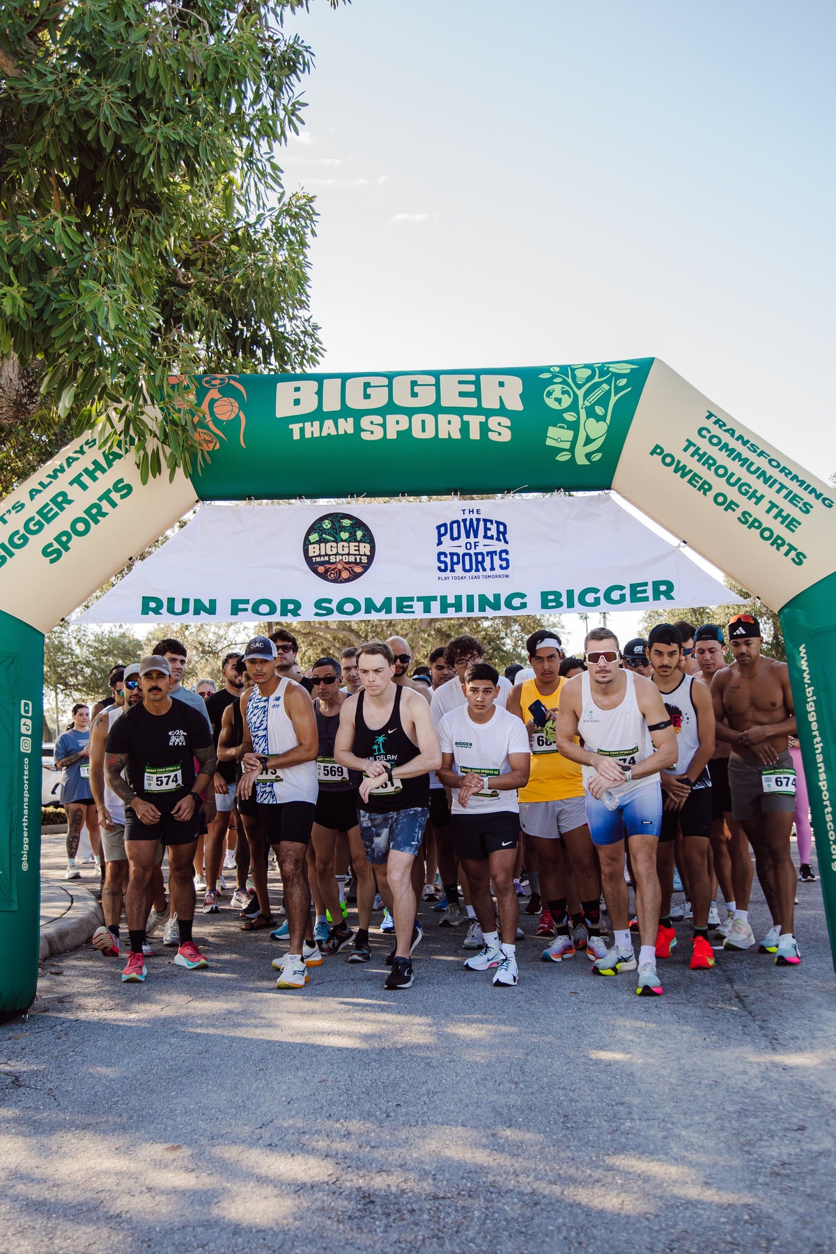 Crowd of runners preparing to start race under a green and white "Bigger Than Sports" inflatable archway