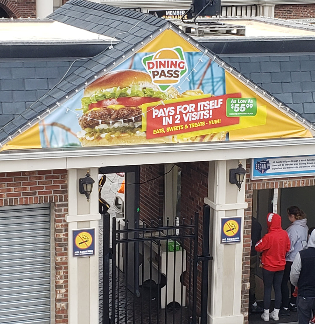 Gable vinyl banner promoting a Six Flags Dining Pass with a large burger image and pricing details, mounted above the entrance to a brick building.