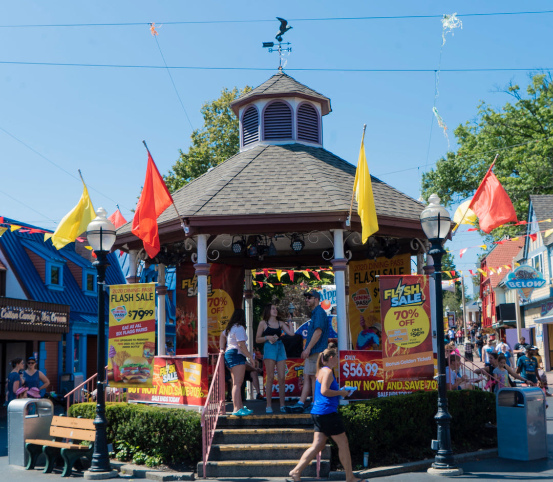Promotional vinyl banners for Six Flags Flash Sale displayed on gazebo with bold text advertising discounts, surrounded by visitors in a vibrant theme park plaza.