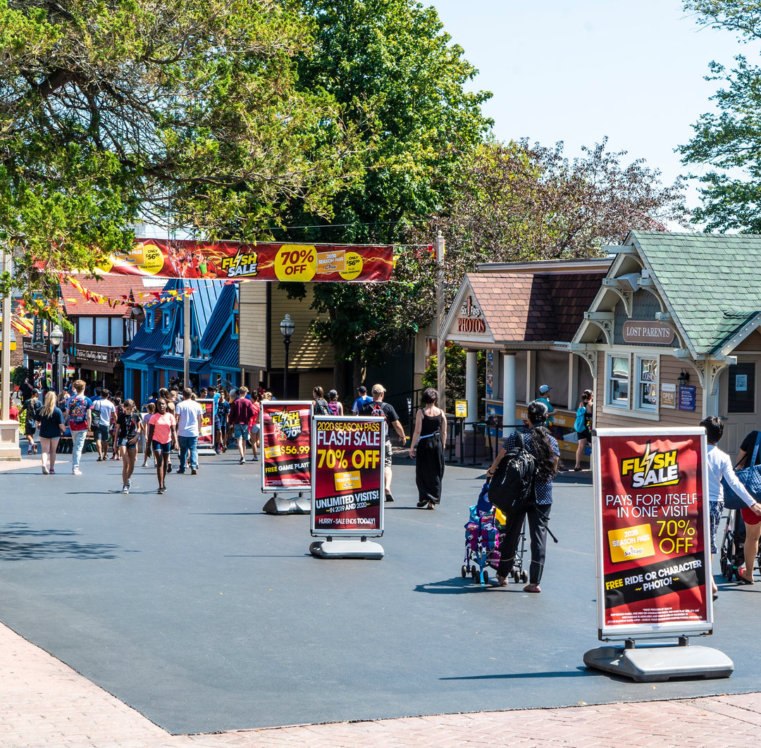 Colorful vinyl banner for Six Flags Flash Sale stretched above main walkway with signage offering 70% off, surrounded by visitors in theme park setting.