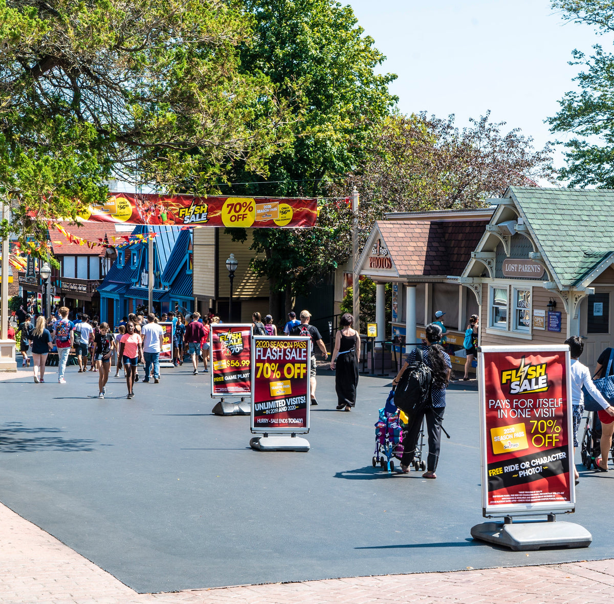 Colorful vinyl banner for Six Flags Flash Sale stretched above main walkway with signage offering 70% off, surrounded by visitors in theme park setting.