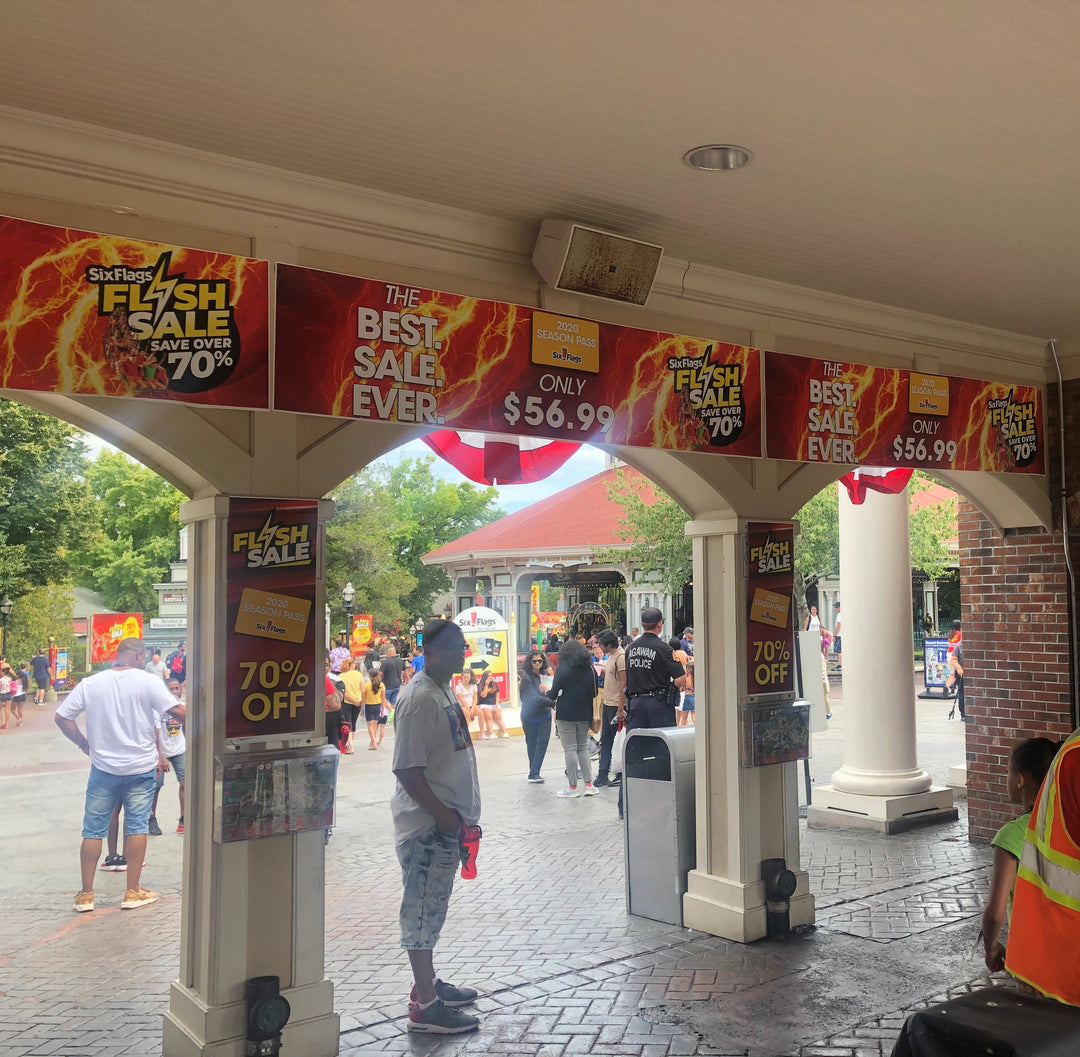 Vinyl banners promoting Six Flags Flash Sale with bold red and yellow graphics offering 70% off season passes displayed around park entrance.