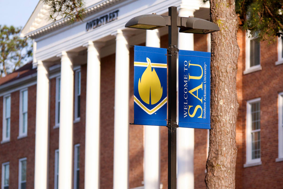 Southern Arkansas University blue light pole banners side by side, with logo emblem on one side and a welcome greeting on the other side, with the campus on the background.