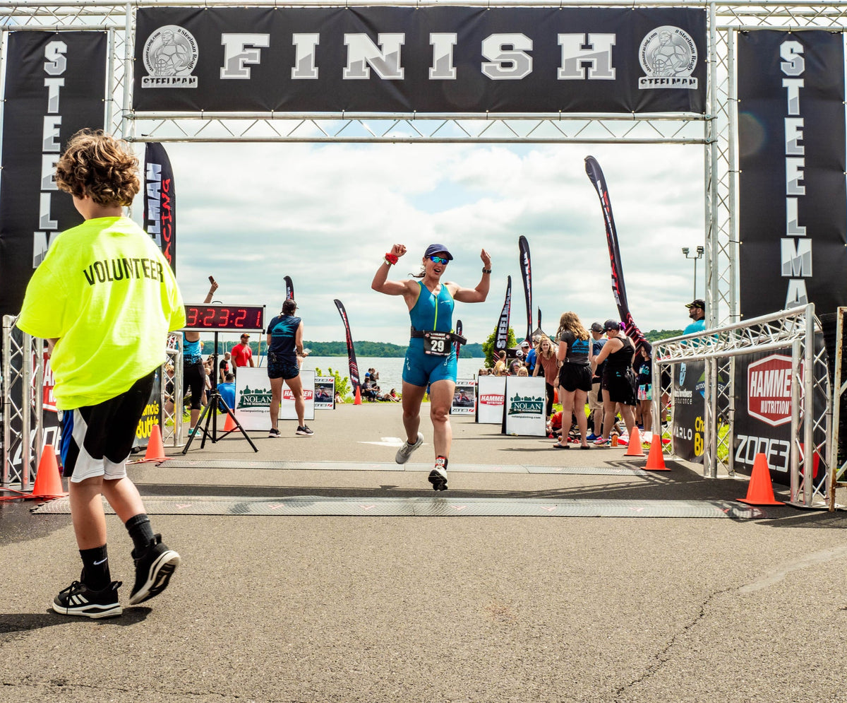 Triathlete finishing race under Steelman Racing branded finish line banner at outdoor event
