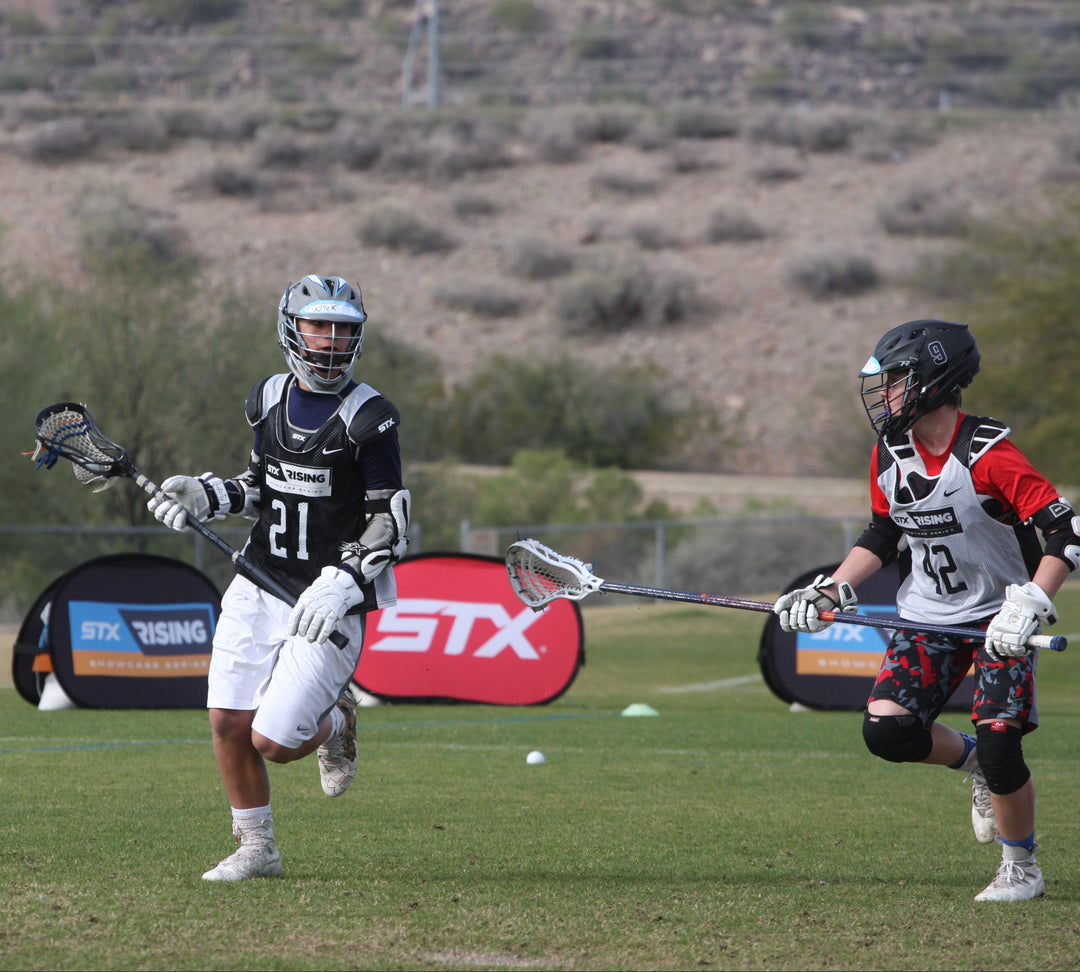 Two youth lacrosse players in action with STX Rising banners in the background on a grassy field