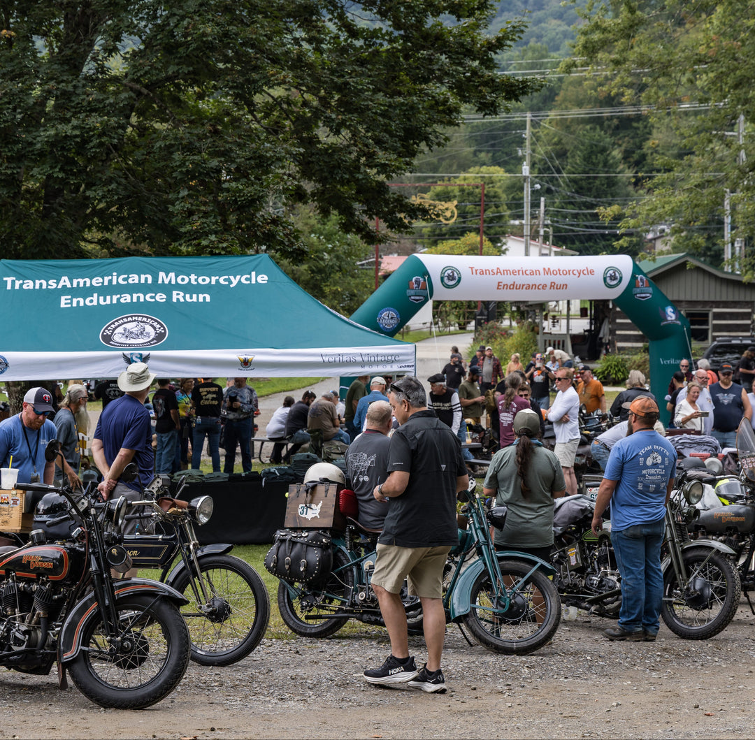 Crowd gathered around vintage motorcycles beneath TransAmerican Motorcycle Endurance Run tent and arch
