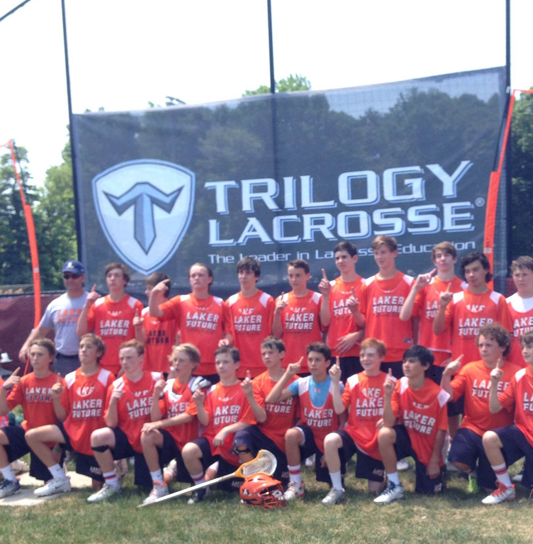 Youth lacrosse team posing for a photo in front of a Trilogy Lacrosse banner on a sunny day