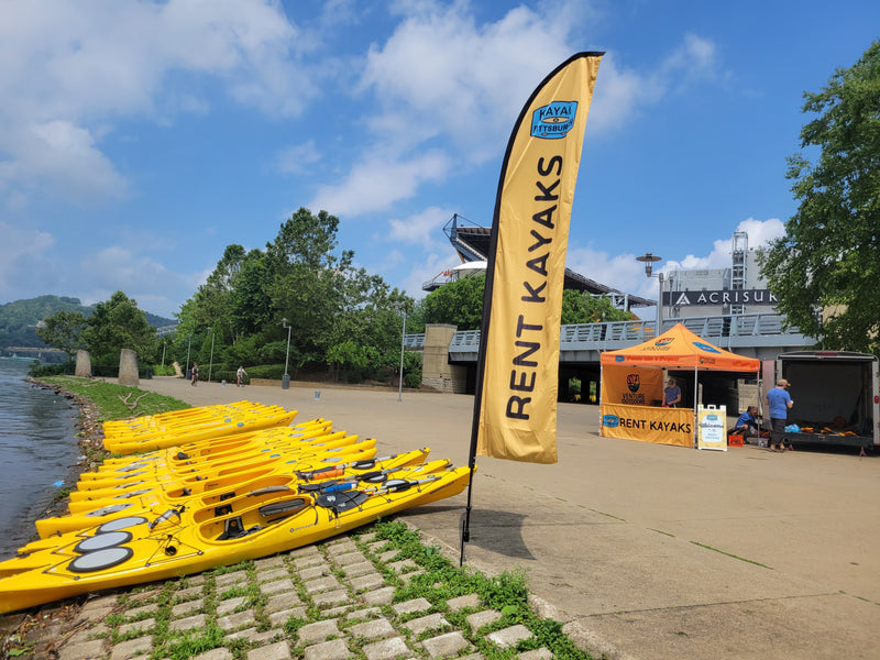 Outdoor feather flag displaying Rent Kayak alongside a vibrant collection of kayaks ready for use